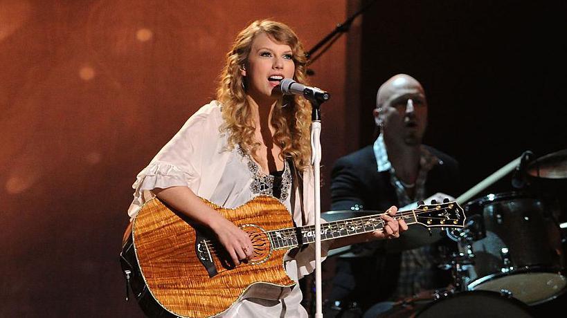 Taylor Swift singing into a microphone on stage during her Speak Now tour, holding a brown guitar.