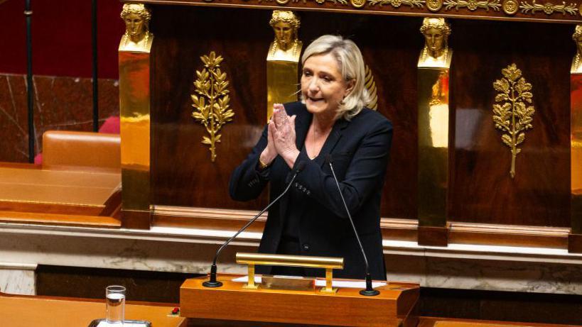 Marine Le Pen, France's far-right National Rally leader, addresses the National Assembly wearing a dark-coloured jacket and holding her hands together