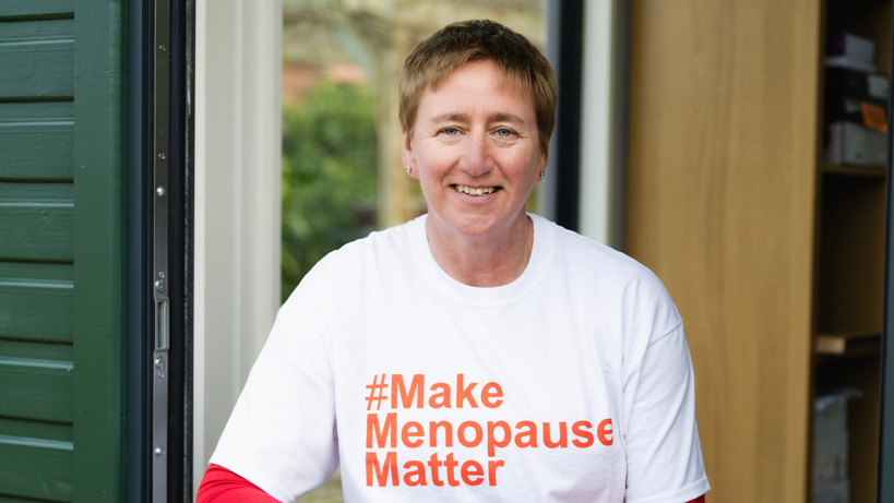 Diane Danzebrink in a doorway smiling at the camera. She wears a white T-shirt with the words '#Make Menopause Matter' written in orange letters in the centre. 