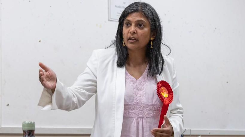 Rupa Huq, wearing a white blazer, which has a red Labour rosette attached to it, giving a speech