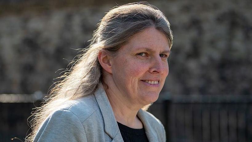 A woman in her late 40s or 50s looks to the side towards the camera while walking. She wears a black top and grey blazer. She has long hair which is light brown and slightly greying.