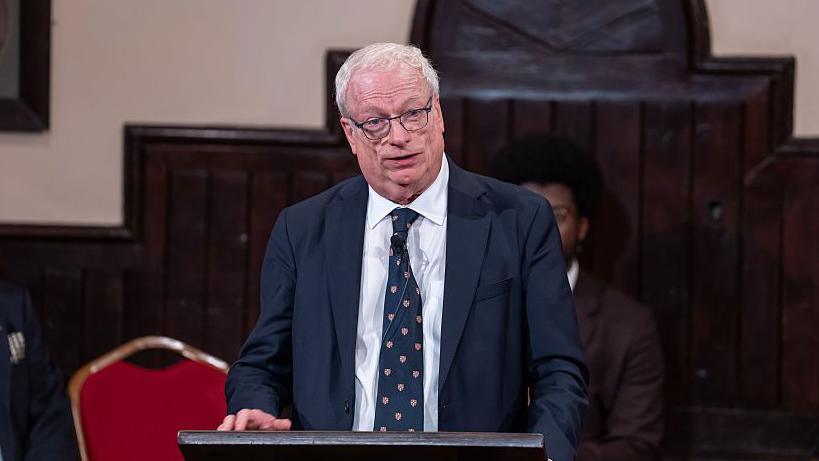 Lord Chris Smith wearing a navy blue suit and white shirt with matching navy blue tie with the Cambridge University crest. He has white hair and black rimmed glasses. He is standing behind a podium.