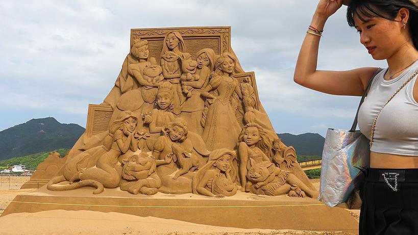 A woman adjusting her sunglasses in front of a sand art pyramid of disney princesses