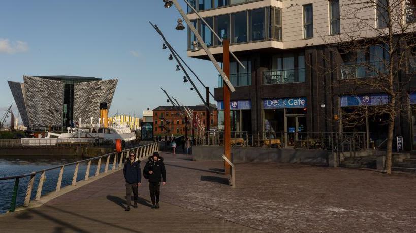 The grey titanic building overlooking a white docked ship in the distance against a blue sky. Then what looks like residential buildings above a blue cafe which is entitled the 'DOCK Cafe'. By the harbour there are two people walking side by side in black with hats on.