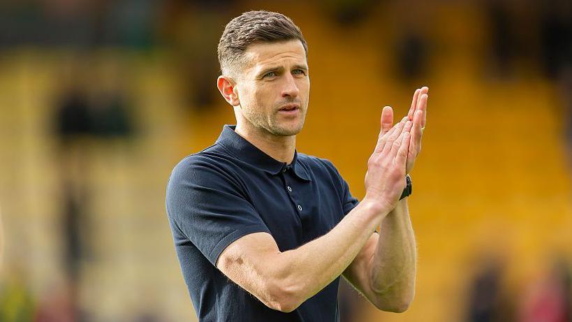 Portsmouth boss John Mousinho in a blue short-sleeved polo top applauds fans after a game at Norwich City