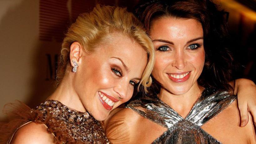 Kylie and Dannii Minogue smiling at the cameras. They are both wearing sparkly tops. Kylie has her arm around Dannii