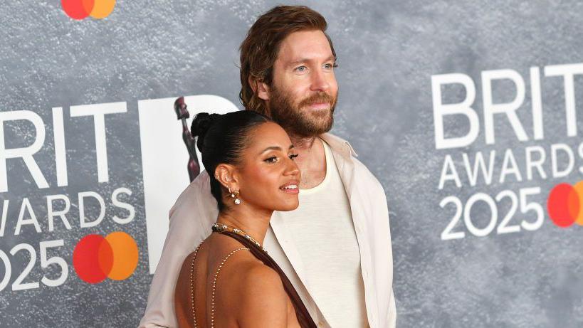 Vick Hope and Calvin Harris pose on the red carpet of the 2025 Brit Awards. He is a head taller than her, and has his arm around her as they both smile. He wears a stylish white shirts over a cream t-shirt and she wears a backless dress with her hair scraped back in a bun.