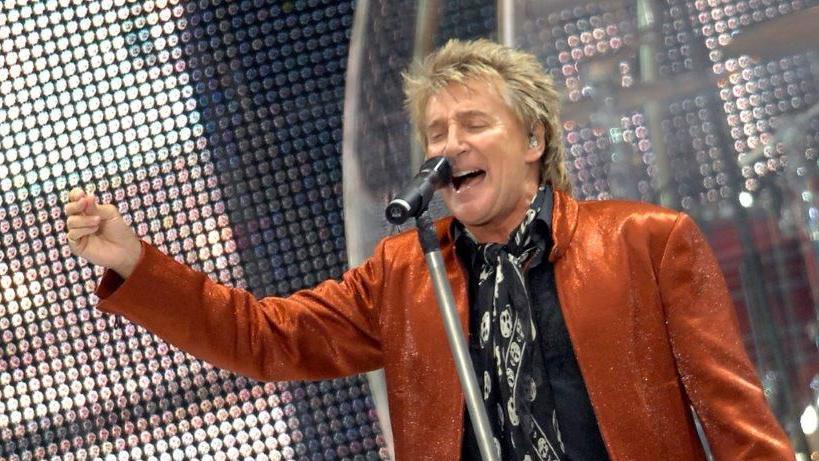Rod Stewart performs at Portman Road on stage. He is wearing an orange suit jacket and a black shirt. He has one arm raised as he sings into a microphone.