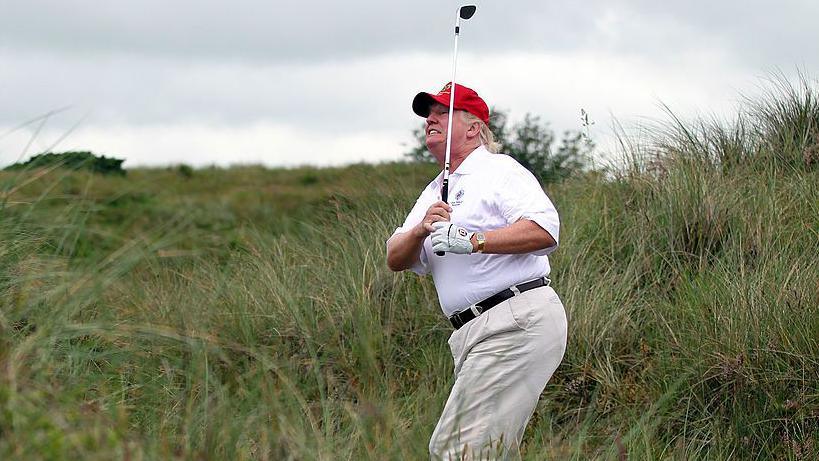 Donald Trump in red golf hat and white t-shirt and trousers hits a golf shot from the rough at his Balmedie course