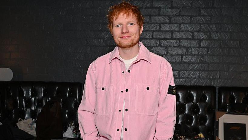 Ed Sheeran standing smiling. He is wearing a pink jacket with a white t-shirt under it.