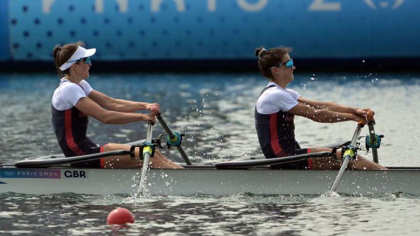 Two women wearing Olympic kits rowing in a white boat with the Paris 2024 logo visible behind them.