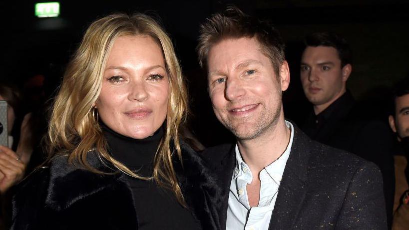 A woman with long blonde hair in a centre parting, wearing a black polo neck and fur jacket smiles next to a man in a flecked black jacket and white shirt. The man has spiky blonde hair.