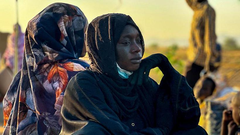 A side image of a woman in a dark headscarf and outfit looking to the right-hand side of the picture. She is sitting on the ground - another woman in a patterned scarf is behind her, she is facing away from the camera.