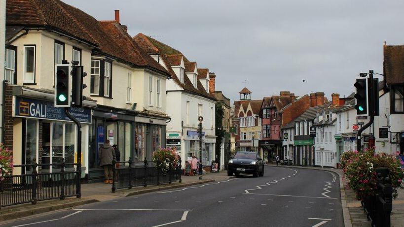 A general view of the High Street in Great Dunmow. It is lined with various shops and businesses. A set of traffic lights are on green, although there is only one car visible and it is parked partially on the pavement. It is an overcast day.