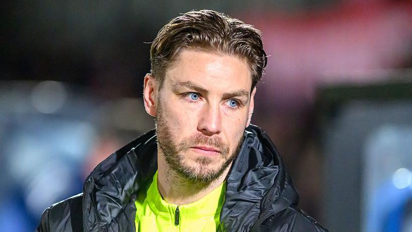 Sam Foley, wearing an unzipped padded coat on top of a fluorescent yellow training top, looks to his left in a pensive manner 