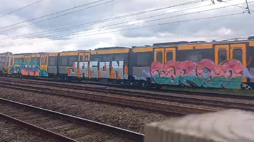 Three metro carriages on a rail sit with graffitti on each, the artwork stretches from the bottom edge of the carriage up to part way through the windows of the trains. The middle carriage has the name Jason painted in silver with fire around the edges while the other two have illegible words coloured in pinks, greens, blues and oranges.