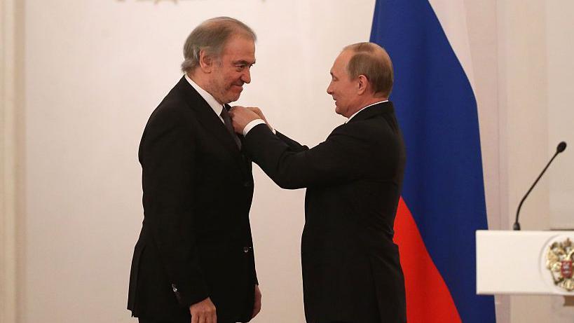 Two men in dark suits stand against a backdrop of a Russian white-blue-and-red tricolor flag, as the man on the right puts his hands on the other man's jacket