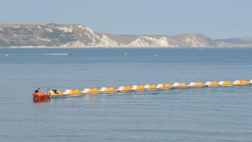 A line of yellow pedalos being towed by an orange rhib boat in Weymouth Bay. the sea is calm and the chalk cliffs of the bay are rising up in the distance.