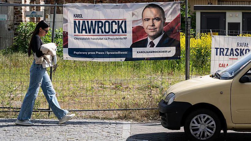 A woman walks past a fence and a campaign banner for Karol Nawrocki