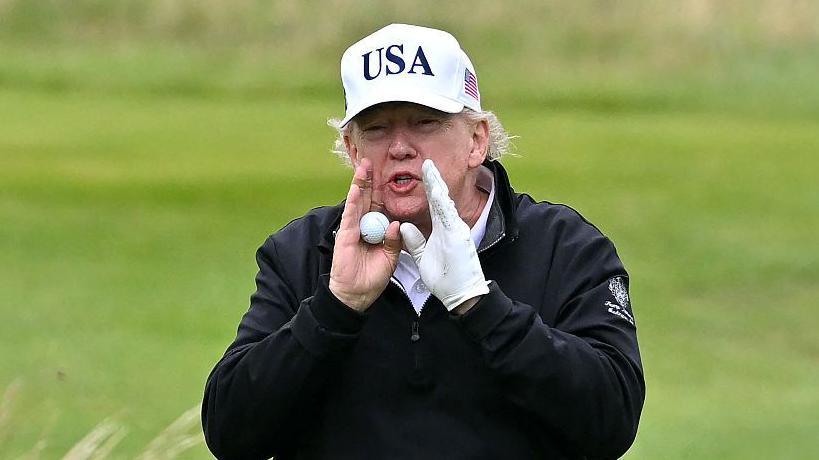 President Trump wearing a white cap with USA on it and a black golf jacket. He has his hands to his mouth as he shouts. He is holding a golf ball in one hand and has a golf glove on the other