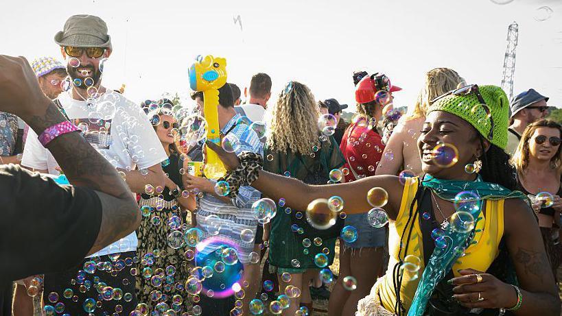 A woman is dancing in bubbles. She is wearing a green hat and yellow top. She is standing in a crowd of people.