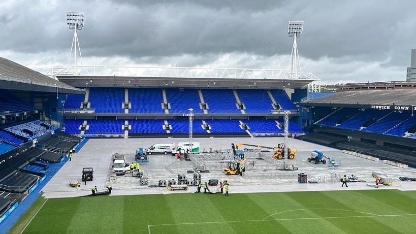 This picture has been taken from high up in one of the stadium's stands behind a goal. It shows vans and heavy machinery vehicles parked around a metal structure in the middle of the pitch, which is partly-covered by white boards. Some of the playing surface is exposed.