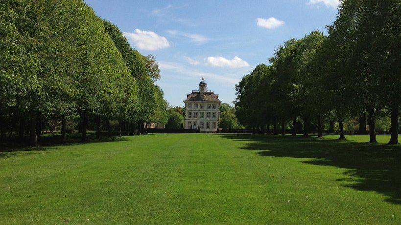 A large three-storey  country house as viewed from the end of a large lawn, rows of trees either side.