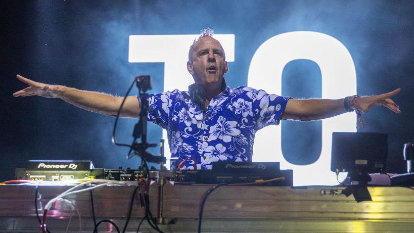 Fatboy Slim performing at an music festival. He is standing behind DJ decks with a large screen behind him. He has his arms stretched to the side and he is wearing a blue shirt with white flowers on it.