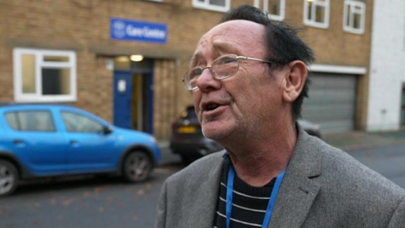 Bill West speaking to a reporter. He is wearing thin-rimmed glasses, an overcoat and a blue lanyard. There is a blue car behind him. 