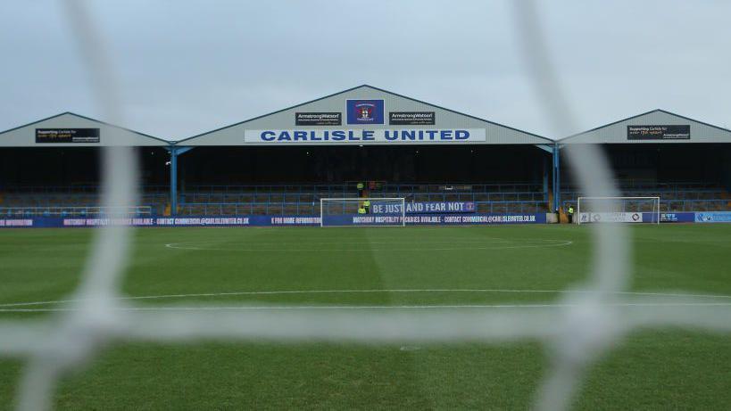 A stand and pitch at Carlisle United ground Brunton Park, pictured through the goal net so the net string is blurry. The words Carlisle United are written on the front of one of the stand roof's peaks.