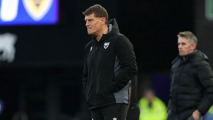 Craig Short watches from the touchline, dressed in a black coat and black tracksuit bottoms, with his hands in his pockets