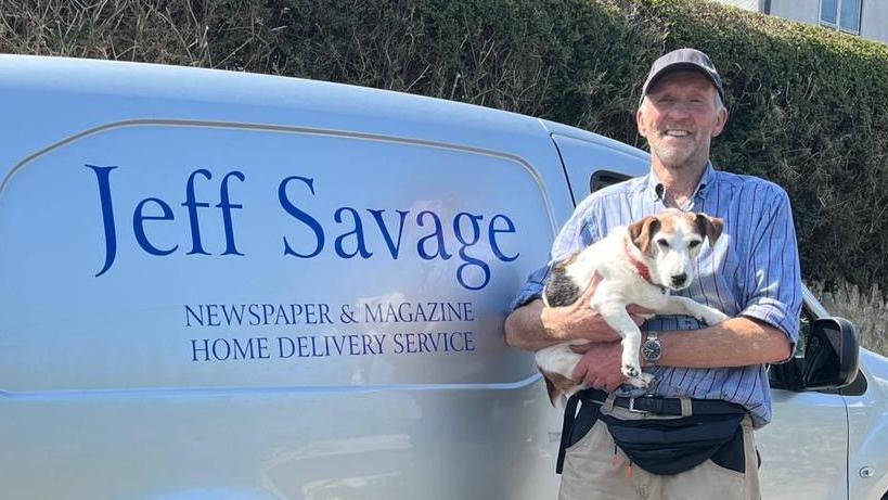 White van with a big blue sign saying "Jeff Savage - Newspaper and Magazine Home delivery service" on the side of it. There's a man in a a blue stripey shirt, with a small dog in his hands.