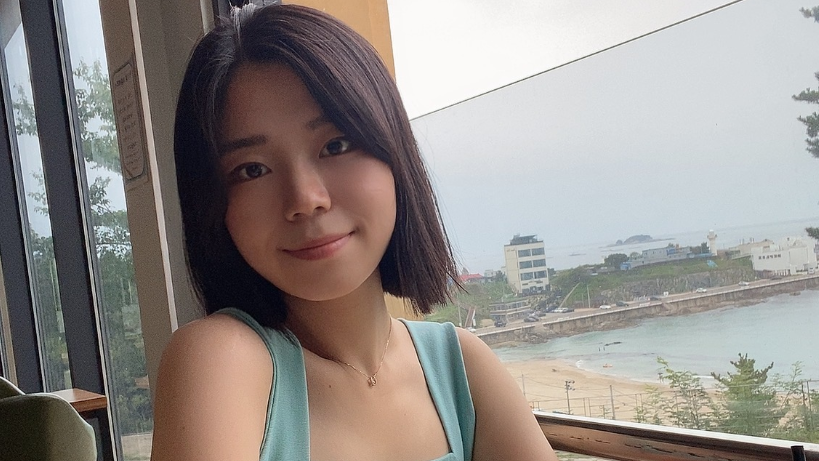 Kim Mi-rim, who has a short dark brown bob and wears a blue vest top, sits in front of a window which looks out onto a seafront 