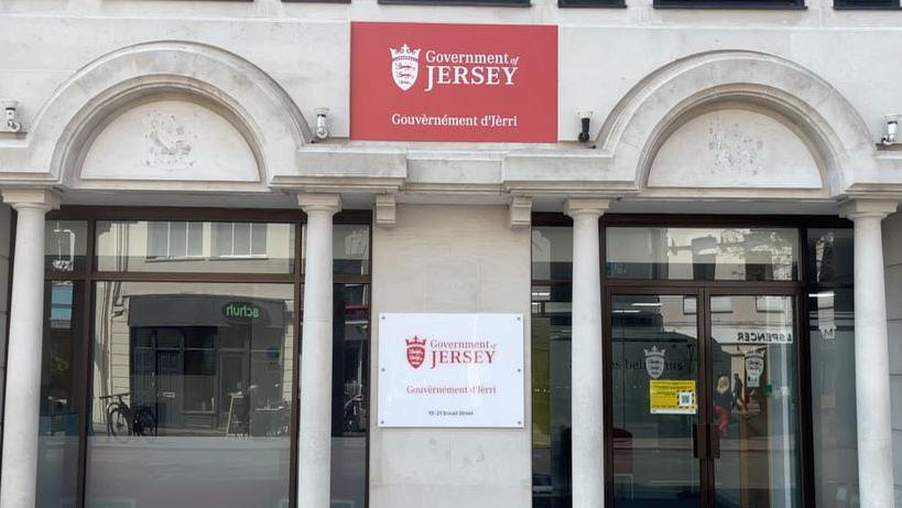 The Government of Jersey building, one red sign in the centre of two pillared windows, in the between lower down a white sign saying the same