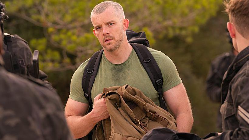 Russell Tovey is in character as Barclay in The War Between. He is outside, stood in front of a backdrop of green trees. He is broad, wearing a green t-shirt with black rucksack straps over his shoulders. He is holding a brown bag and surrounded by what looks like a group of military staff in uniform.