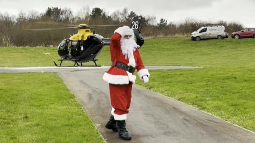 Father Christmas walking away from the helicopter down a tarmac path, with green grass either side of him. The sky is grey and cloudy, but his festive outfit stands out against the backdrop.