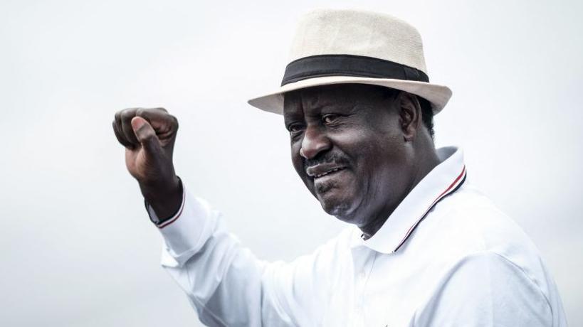 Raila Odinga wearing a hat and white shirt raises his fist to supporters