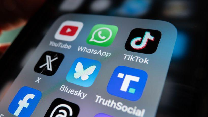 An iPhone screen is lit up and shows an open folder with various social media logo apps including YouTube, WhatsApp, TikTok, X, Bluesky, TruthSocial and Facebook