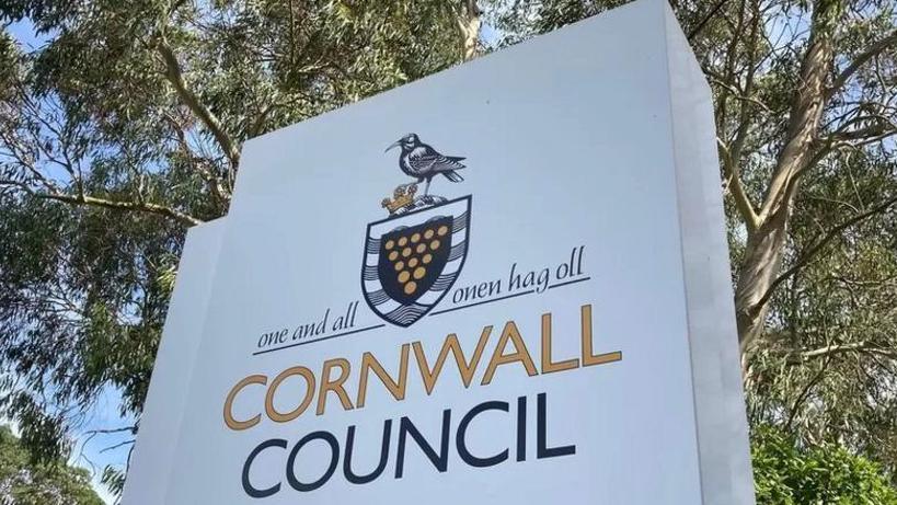 A white sign which says Cornwall Council in yellow and black writing, with a shield emblem with a bird above it and a motto that says "one and all" in both English and Cornish