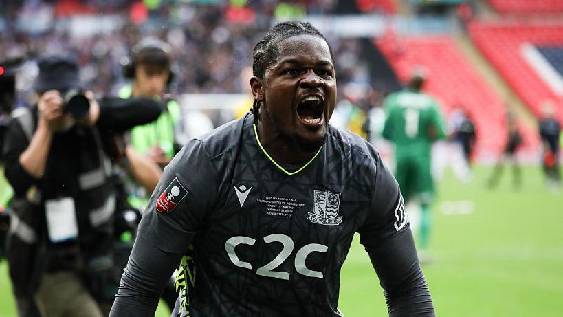 Goalkeeper Andeng Ndi yells into camera at side of pitch at Wembley, with cameraman in soft focus taking shots from behind