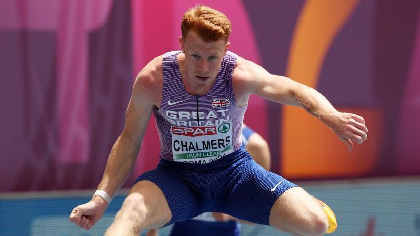 Alastair Chalmers competing in the hurdles. He is wearing a Great Britain vest and blue shorts.