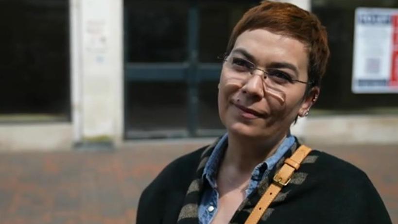 Rodica White, a woman wearing a blue shirt and cardigan with a bag over her shoulder standing on a street