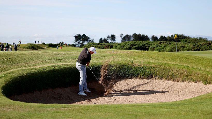 A wide shot of a golfer wearing a dark polo shirt, light grey trousers and a white cap chipping a golf ball out of a bunker. Sand can be seen spraying up where his club and the ball made contact and the ball has lifted just out of the bunker. In the distance the yellow flag marking the hole can be seen as well as some scrubland and, further up the image, the blue sky.