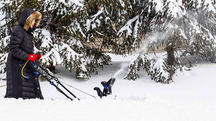 A woman pushes a snow removing machine in deep snow. The snow is spraying out of the machine and pine trees are behind her. She is wearing a black long coat and red gloves.