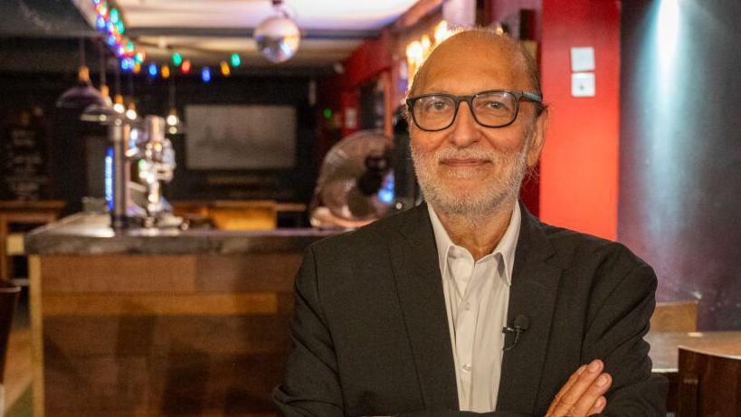 Man wearing glasses and a shirt and jacket stands inside an empty bar with his arms crossed. He looks directly at the camera. A wooden bar with twinkly-coloured lights above it, down lights and beer pups are to the left and behind him. A small disco ball hangs from the ceiling of the bar area.
