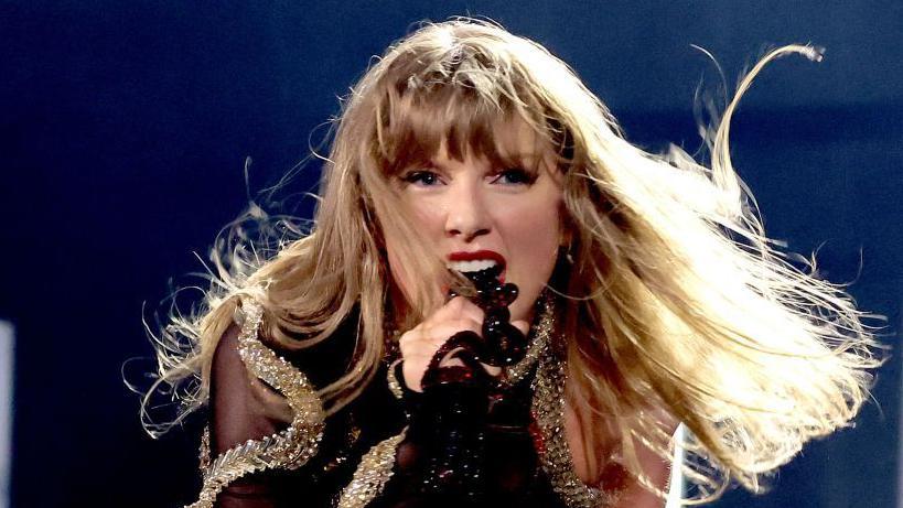 Taylor Swift performs on stage in December last year in Vancouver, Canada, as part of her Eras tour. She is singing passionately into the microphone and is wearing a black and gold sequinned outfit.