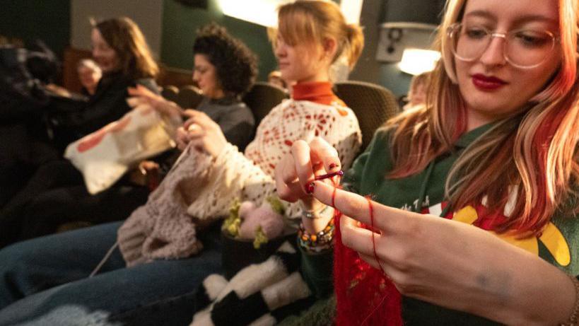 Women sit in a cinema and knit and crochet.