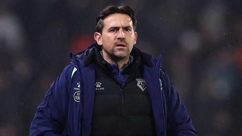 A concerned looking Charlie Daniels in a dark blue Watford overcoat and black tracksuit top with the club badge on them