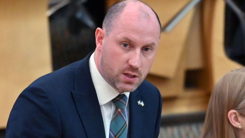 Neil Gray, a bald man wearing a navy suit with a tartan tie. He is standing in the Scottish parliament.