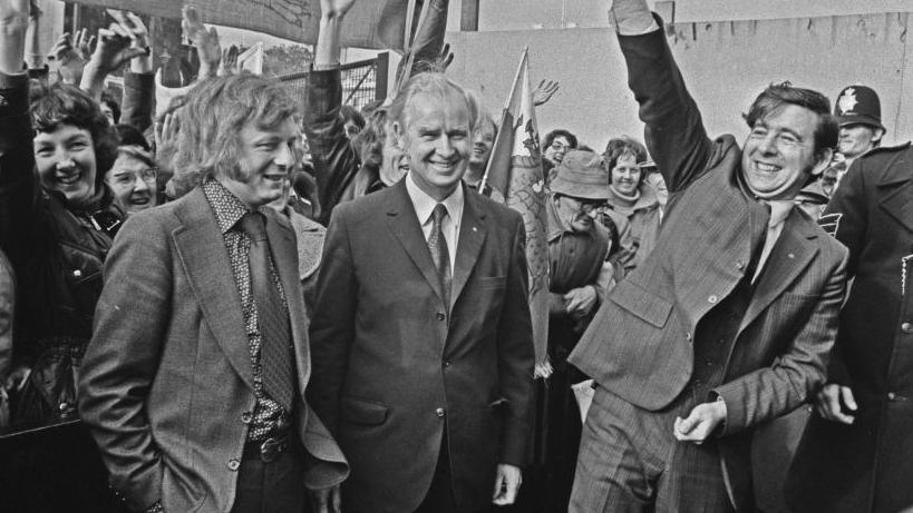 A black and white photograph of Dafydd Elis-Thomas, Gwynfor Evans and Dafydd Wigley surrounded by supporters at the House of Commons in 1974